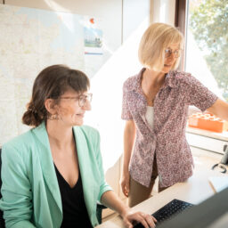 Zwei Frauen im Büro schauen gemeinsam auf einen Monitor, eine trägt ein mintgrünes Sakko, die andere steht neben ihr