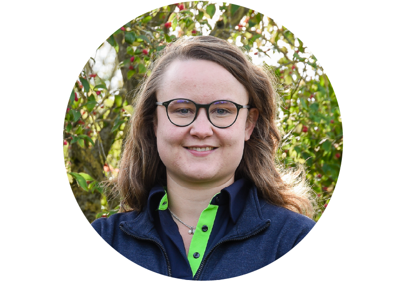 Frau mit brille und schulterlangen braunen Haaren steht in KVG-Dienstkleidung vor einem Strauch, lächelt freundlich