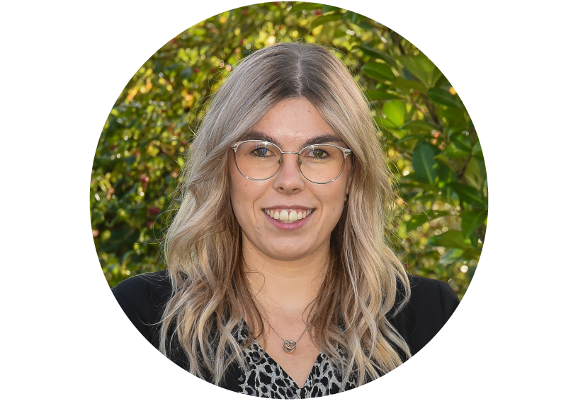 Frau mit langen, hellblonden Haaren und großen Brillen steht vor grünem Laub, trägt schwarzes Oberteil mit Muster, lächelt offen