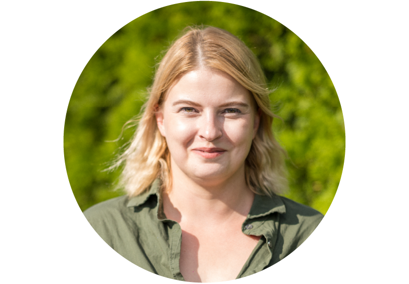 Frau mit schulterlangen blonden Haaren steht vor sonnigem Grün, trägt ein offenes olivgrünes Hemd, schaut direkt in die Kamera