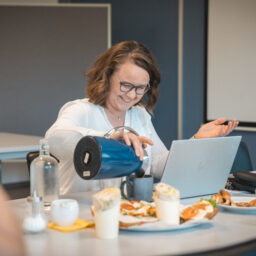 Frau gießt Kaffee aus Thermoskanne an Tisch mit Pizza und Getränken, im Hintergrund Laptop und Büroambiente