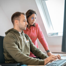Junger Mann in grünem Kapuzenpulli arbeitet mit Kollegin im pinken Pullover an Computer in modernem Büro